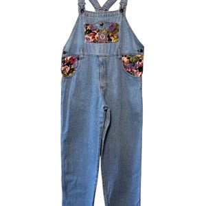 Può includere: Salopette in denim blu chiaro con dettagli floreali sulla pettorina e sulle tasche. La salopette ha spalline regolabili, chiusura con bottoni e un design classico. La stampa floreale presenta una varietà di colori.