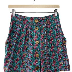 May include: A floral print skirt with a button-up front. The skirt features a vibrant pattern of red, purple, and yellow flowers on a teal background. It has large wooden buttons and two pockets. The skirt is hanging on a wooden hanger.