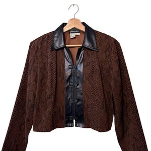 Peut inclure: Veste marron à motif peau de serpent avec col et panneau avant en cuir noir. La veste a des manches longues et une fermeture à crochet et œillet. La veste est suspendue à un cintre en bois.