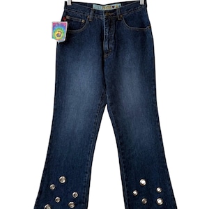 May include: Dark wash denim jeans with a flared leg design. The jeans feature silver grommet details along the bottom hem. A tag with a tie-dye design and the word "mudd" is attached to the jeans.