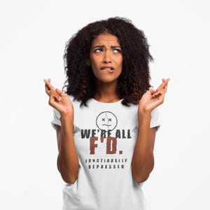 May include: A white short-sleeved t-shirt featuring a graphic of a sad face with X-shaped eyes and the text WE'RE ALL F'D. FUNCTIONALLY DEPRESSED printed in black and brown block lettering on the front center of the garment.