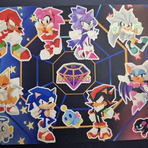 Puede incluir: Una hoja de pegatinas con personajes de la franquicia Sonic the Hedgehog. Incluye a Sonic, Tails, Knuckles, Amy, Blaze, Silver, Shadow y Rouge, sobre un fondo azul y dorado.