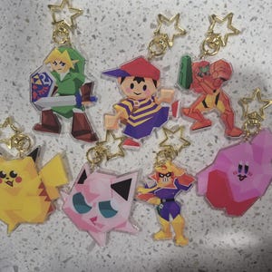 Puede incluir: Una colección de llaveros acrílicos coloridos con varios personajes de videojuegos. Los llaveros incluyen personajes como Link, Pikachu y Kirby, cada uno con un cierre dorado en forma de estrella.
