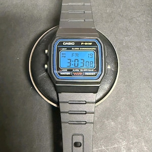 May include: A black Casio F-91W digital watch with a blue-toned display showing the time, day, and date. The watch has a black strap and a rectangular face with the Casio and F-91W model names. The display reads 3:03:08.