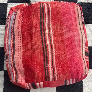 May include: A square, red pouffe with a striped pattern in shades of red, pink, black, and white. The woven fabric has a textured appearance. The pouffe is a decorative accent for living rooms or bedrooms.