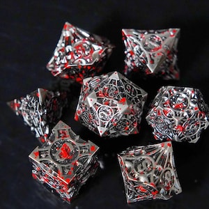 May include: A set of seven polyhedral dice featuring a metallic silver finish with intricate openwork lattice designs and irregular bright red splatters across the surfaces. Each die displays clear black numerals etched into the metal framework.