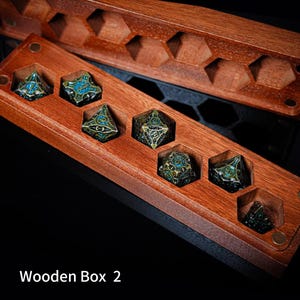 May include: A rectangular wooden box with hexagonal cutouts containing six multi-faceted metal dice. The dice feature an intricate dark bronze finish with blue enamel accents. The box has a matching wooden lid with hexagonal patterns. Text reads Wooden Box 2.