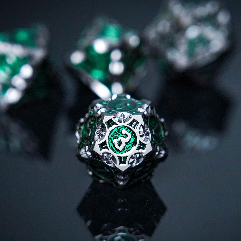 May include: A twenty-sided die featuring a metallic silver frame with intricate cutouts and a vibrant emerald green inlay. The numbers are embossed in a stylised font within the green sections, creating a sharp contrast against the polished silver metal.