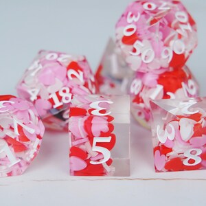 May include: A set of polyhedral dice made of clear resin filled with small red, pink, and white heart-shaped confetti. Each die features bold white numbers printed on the exterior surfaces, including a d20, a d12, a d10, a d8, and a d4.