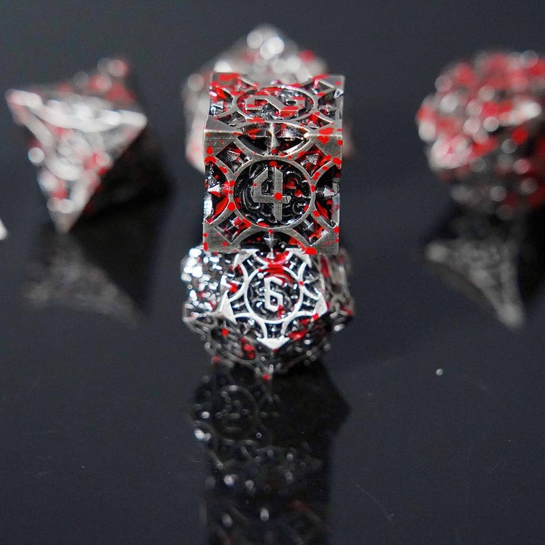 May include: A set of metallic polyhedral dice featuring an intricate, openwork cage design with a dark silver finish and splattered red paint accents. The dice display clear black numerals, with a four-sided die stacked atop a six-sided die.