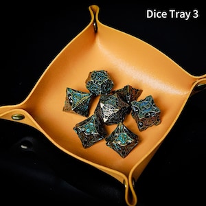 May include: A tan, square-shaped folding dice tray with metal snap fasteners at each corner, containing seven intricate, dark metallic polyhedral dice with blue glitter-filled filigree patterns. The text Dice Tray 3 is visible.