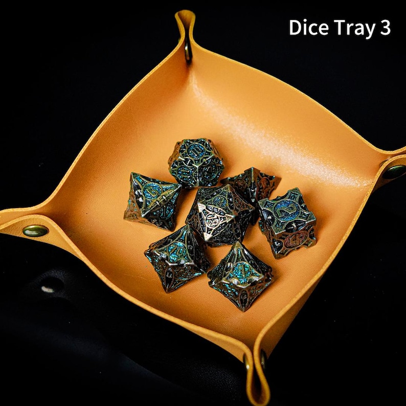 May include: A tan, square-shaped folding dice tray with metal snap fasteners at each corner, containing seven intricate, dark metallic polyhedral dice with blue glitter-filled filigree patterns. The text Dice Tray 3 is visible.