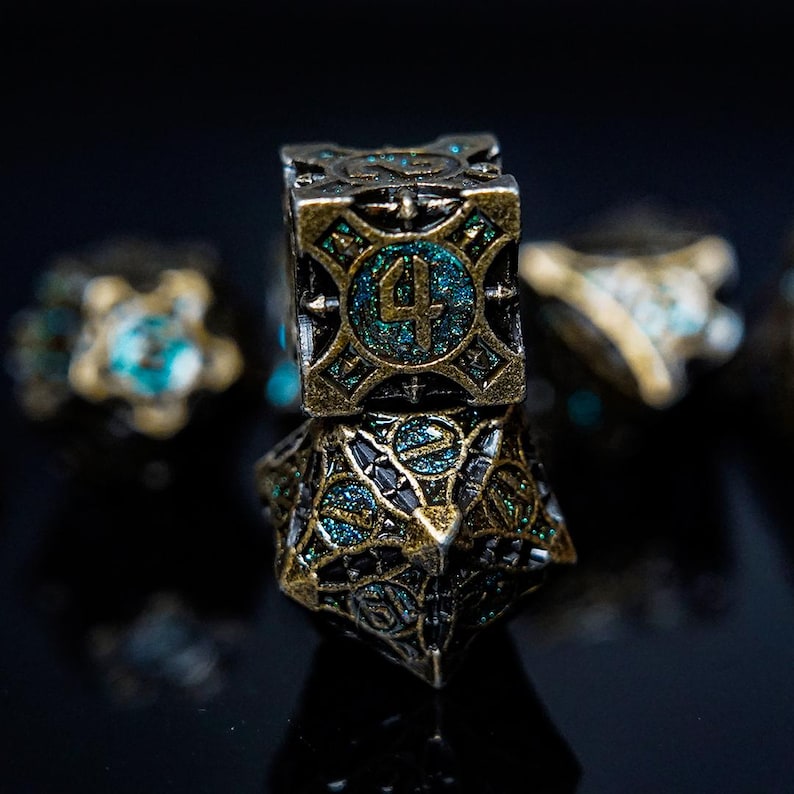 May include: A set of metal polyhedral dice featuring an antique brass finish with intricate, raised geometric detailing and recessed teal glitter enamel inlays. The top cube-shaped die clearly displays the number four in a bold, embossed font.