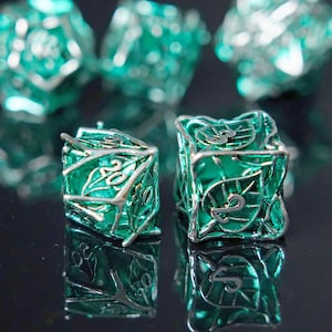 Puede incluir: Primer plano de varios dados de color verde azulado con detalles plateados. Los dados tienen un diseño de hojas y están numerados. Los dados están sobre una superficie reflectante, con algunos dados desenfocados en el fondo.