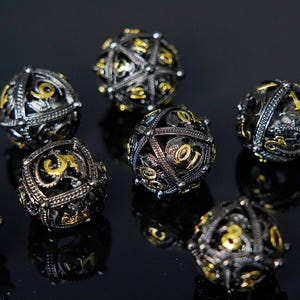May include: A collection of spherical dice with a black and gold colour scheme. The dice have an ornate metal design with gold-coloured numbers. These dice are likely used for tabletop gaming, with each die showing a different number.