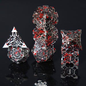 May include: A set of metal polyhedral dice featuring an intricate, openwork cage design in a dark silver finish with bright red paint splatters. The dice display clear numerical engravings and sharp geometric edges, arranged in three distinct stacks.