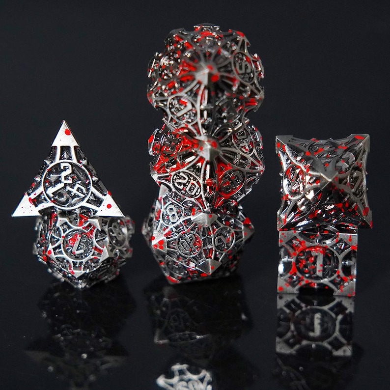 May include: A set of metal polyhedral dice featuring an intricate, openwork cage design in a dark silver finish with bright red paint splatters. The dice display clear numerical engravings and sharp geometric edges, arranged in three distinct stacks.