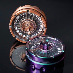 Puede incluir: Dos dados giratorios de metal con esferas numeradas. Uno es de color cobre, el otro es morado con acentos iridiscentes. Cada giratorio tiene una ventana circular transparente que muestra los números del 1 al 20. Los dados están sobre una superficie oscura y reflectante.