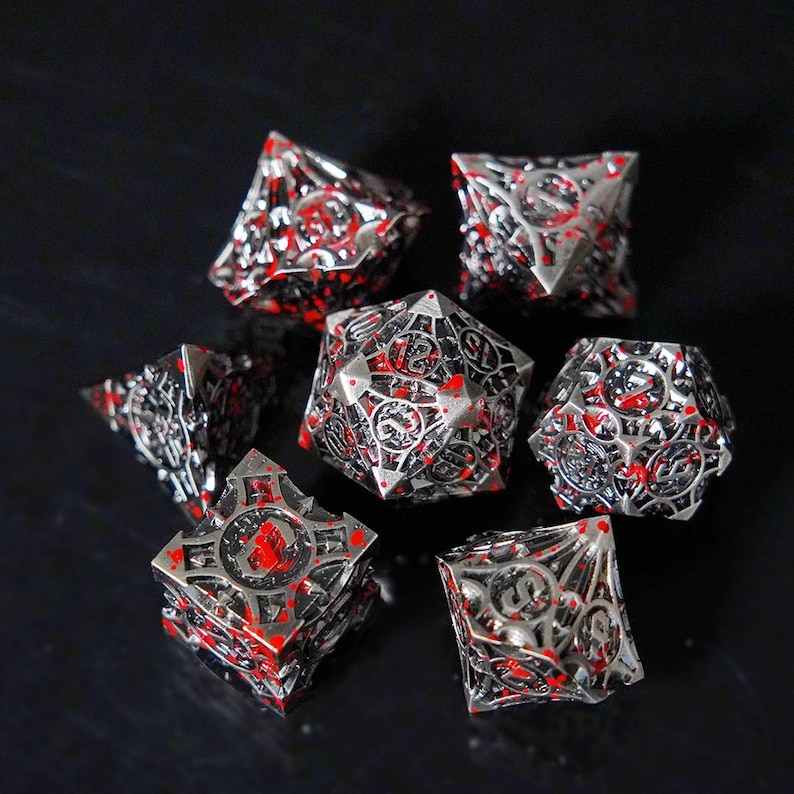 May include: A set of seven polyhedral dice featuring a metallic silver finish with intricate openwork lattice designs and irregular bright red splatters across the surfaces. Each die displays clear black numerals etched into the metal framework.