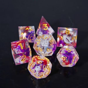 Puede incluir: Juego de siete dados poliédricos con base de resina transparente, con remolinos de pigmentos morados, rojos y blancos. Cada dado tiene números dorados. Diseñado para juegos de rol de mesa.
