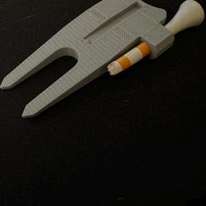 Puede incluir: Una herramienta gris impresa en 3D para reparar divots de golf con un extremo en forma de horquilla y un mango. Un objeto a rayas blancas y naranjas se inserta en la herramienta. La herramienta está sobre una superficie negra.