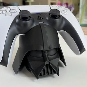 May include: A white and black PlayStation 5 controller resting on a black Darth Vader-themed charging stand. The controller has black joysticks and buttons. The stand is shaped like Darth Vader's helmet.