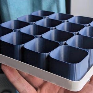 May include: A tray with sixteen dark blue square containers. The containers are arranged in a grid pattern and have rounded corners. The tray has a white border and is held by a hand.