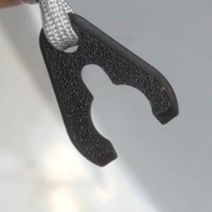 Könnte beinhalten: Ein schwarzer, strukturierter Kunststoffclip mit geschwungenem Design und einem Loch in der Mitte, befestigt an einer grau-weißen geflochtenen Kordel. Der Clip hat eine matte Oberfläche und scheint ein funktionelles Element zu sein.