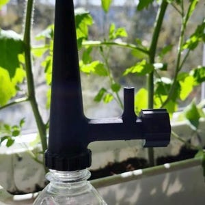 May include: A black plastic plant watering device attached to a clear plastic bottle. The device has a long, tapered spout and a side nozzle. The background features green plants in a white planter, suggesting indoor gardening.