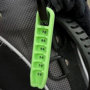 May include: A lime green golf club distance marker with black numbers indicating distances from 20 to 70 yards. The marker is attached to a black clip and is hanging on a golf bag.