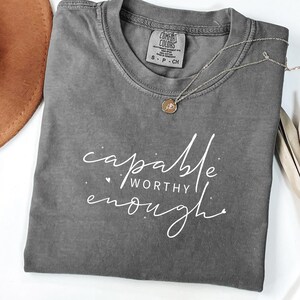 Könnte beinhalten: Ein anthrazitfarbenes Kurzarm-T-Shirt mit Rundhalsausschnitt und einem Comfort Colors Markenetikett. Auf der Vorderseite befindet sich ein weißer Schriftzug "capable WORTHY enough" in einer Mischung aus Schreib- und Blockschrift, verziert mit kleinen Punkten und einem Herz.