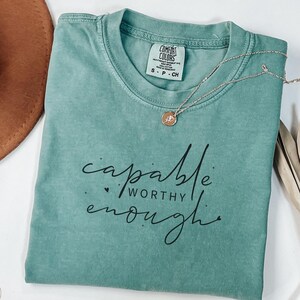 Könnte beinhalten: Ein salbeigrünes Kurzarm-T-Shirt mit Rundhalsausschnitt und einem Comfort Colors Markenetikett. Das Shirt ist mit dem schwarzen Schriftzug capable WORTHY enough bedruckt, verziert mit kleinen Herz- und Punktesymbolen, sowie einer dünnen goldfarbenen Kette.