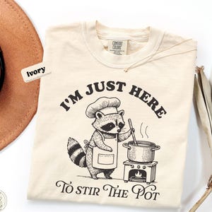Puede incluir: Camiseta color marfil con un gráfico de un mapache cocinero y el texto "I'm Just Here To Stir The Pot". El mapache lleva un gorro de chef y un delantal, removiendo una olla en una estufa. Marca Comfort Colors.