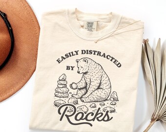 Op de afbeelding: Een crèmekleurig t-shirt met korte mouwen en een donkerbruine afbeelding van een beer die zit en stenen stapelt. De tekst boven de beer luidt "EASILY DISTRACTED BY" en het woord "Rocks" staat eronder in een groot cursief lettertype.