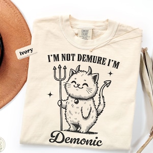 May include: An ivory t-shirt featuring a black graphic of a cat with horns and a tail, holding a trident. The text reads I'M NOT DEMURE I'M Demonic. The shirt has a Comfort Colors label at the neck and is displayed alongside a brown hat and dried leaves.