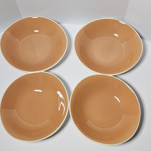 May include: A set of four round ceramic bowls with a smooth, glossy finish in a solid light peach color. Each bowl features a simple, wide-rimmed design with a clean, white edge along the circumference, presenting a uniform and consistent appearance.