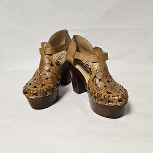 May include: A pair of vintage brown leather platform sandals. The sandals feature floral cutouts, a buckle closure, and a thick wooden heel. The platform measures approximately 2 inches, and the heel is about 4 inches high.