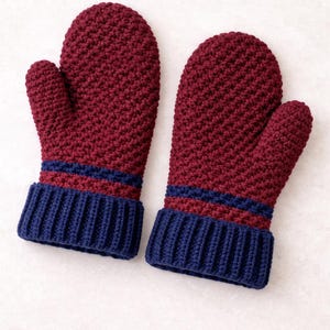 K&ouml;nnte beinhalten: Ein Paar bordeauxfarbene H&auml;kelhandschuhe mit marineblauen Rippb&uuml;ndchen. Die F&auml;ustlinge haben ein strukturiertes Stichmuster und zwei marineblaue Streifen &uuml;ber den B&uuml;ndchen. Warme Winteraccessoires.
