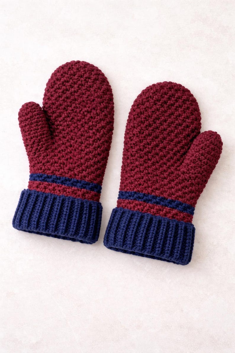 K&ouml;nnte beinhalten: Ein Paar bordeauxfarbene H&auml;kelhandschuhe mit marineblauen Rippb&uuml;ndchen. Die F&auml;ustlinge haben ein strukturiertes Stichmuster und zwei marineblaue Streifen &uuml;ber den B&uuml;ndchen. Warme Winteraccessoires.