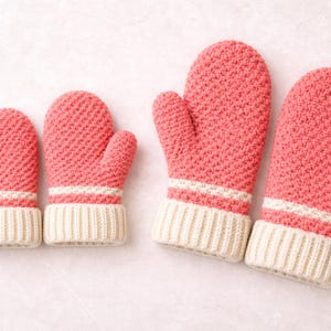 K&ouml;nnte beinhalten: Ein Satz korallenfarbener Strickf&auml;ustlinge mit strukturiertem Muster. Jeder F&auml;ustling hat eine cremefarbene gerippte Manschette und einen wei&szlig;en Streifen. Die F&auml;ustlinge sind paarweise angeordnet und deuten auf verschiedene Gr&ouml;&szlig;en hin.