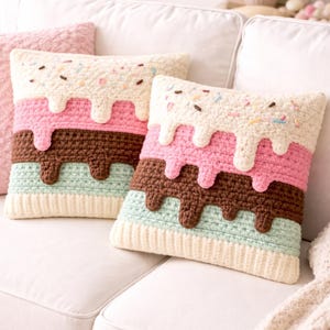 May include: Two square crochet cushions with an ice cream design. The top layer is off-white with sprinkles, followed by pink, brown, and light blue layers. The cushions are on a white sofa.