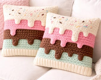 May include: Two square crochet pillows with a layered ice cream design. The top layer is off-white with sprinkles, followed by pink, brown, and light blue layers. The pillows are on a white couch.