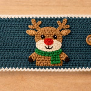 May include: Teal crochet cosy featuring a reindeer design. The reindeer has a red nose, green scarf, and brown antlers. The cosy has a white border and a wooden button closure. A loop is attached for hanging.