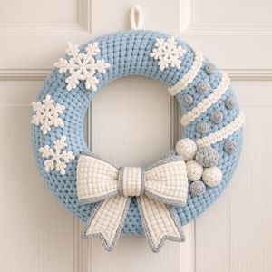 Puede incluir: Una corona de ganchillo azul claro con detalles de copos de nieve blancos y un lazo grande. La corona presenta un diseño texturizado con adornos blancos y grises, ideal para la decoración invernal. La corona mide unos 30 cm de diámetro.