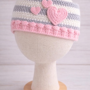 May include: A handmade crochet headband in white, gray, and pink, adorned with three pink hearts. The headband features a striped pattern and a textured pink border, displayed on a mannequin head.