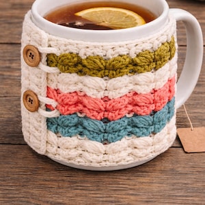 May include: A white ceramic mug filled with tea and a lemon slice, wrapped in a colorful, hand-knitted cozy. The cozy features cream, olive green, coral, and teal stripes, secured with wooden buttons. A tea bag tag hangs from the mug.