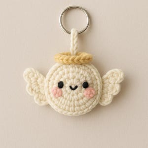 May include: A handmade crochet angel keychain. The angel has a round body, small wings, and a halo. The face features black eyes, a small smile, and pink cheeks. The keychain is attached to a silver metal ring.
