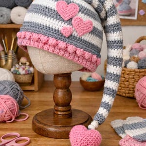 Puede incluir: Un gorro de ganchillo hecho a mano con una larga cola a rayas, exhibido en un soporte de madera. El gorro presenta rayas grises, blancas y rosas, con aplicaciones de corazón rosas y un ribete de pompón rosa. Un corazón rosa a juego está unido a la cola.