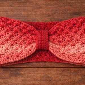 May include: A handmade, red and coral crochet headband with a bow-like design. The headband is made of soft yarn and features a textured stitch pattern. The item is displayed on a wooden surface.