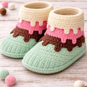 Può includere: Un paio di pantofole all'uncinetto fatte a mano progettate per assomigliare a coni gelato. Le pantofole presentano una base verde menta, uno strato intermedio marrone, uno strato rosa e una parte superiore color crema, con un effetto gocciolante. Le pantofole sono su una superficie di legno chiaro.